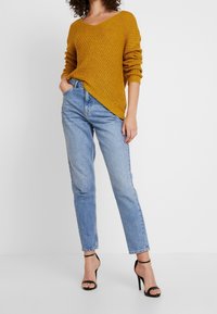 Hořčicově žlutý pletený svetr s výstřihem do V, dlouhými rukávy a žebrovanou strukturou, v kombinaci se světle modrými rovnými džíny a černými sandály na podpatku.