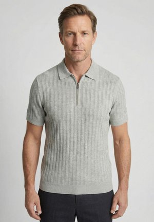 Mann trägt ein hellgraues, kurzärmeliges, geripptes Strick-Poloshirt mit einem halbhohen Reißverschlusskragen und steht vor einem einfarbigen hellen Hintergrund.