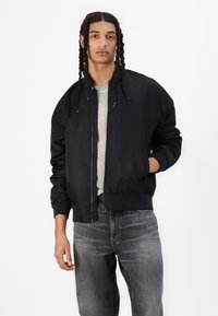 Zwarte bomberjack met een gladde afwerking, ribbelmanchetten en een ritssluiting, gedragen over een grijs ribbeltop en gecombineerd met grijze jeans.