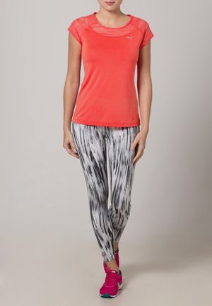 Nainen pukeutunut korallinväriseen lyhythihaiseen treenipaitaan, mustavalkokuosillisiin treenilegginseihin ja kirkkaanpinkoihin urheilukenkiin, seisomassa neutraalia taustaa vasten.