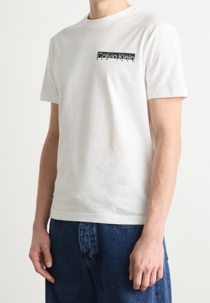 Mężczyzna noszący białą koszulkę Calvin Klein z czarnym logo na piersi, w połączeniu z niebieskimi dżinsami, stoi na jednolitym tle.