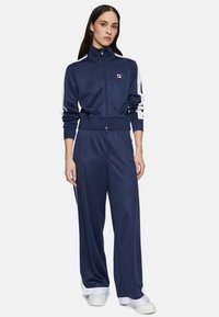 Tuta navy realizzata in tessuto morbido con strisce verticali. Presenta un colletto alto, chiusura con zip e accenti bianchi sui lati. Sneakers bianche completano il look.