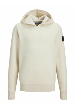 Person iført en sort polstret vest over en beige hættetrøje med lyse, vasket løse jeans og hvide sneakers mod en ensfarvet baggrund.