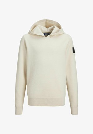 Person iført en sort polstret vest over en beige hættetrøje med lyse, vasket løse jeans og hvide sneakers mod en ensfarvet baggrund.