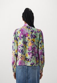 Blouse à manches longues florale dans des tons de violet, vert et jaune. Présente des poignets smockés et un tissu léger et texturé.