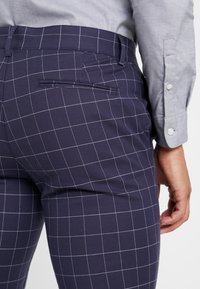 Marinblå rutig byxor med slim fit, med platt front, sidofickor och en enda bakficka. Materialet verkar mjukt och lätt.