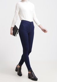 Tmavě modré vysoké džíny jsou zkombinovány s bílým topem s dlouhým rukávem a černými sandály na podpatku s krajkovým vzorem; drží šedý psaníčko.