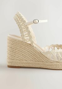 Sandale compensée tissée beige avec boucle de bride à la cheville et détails en corde tressée au talon et sur les parties supérieures.