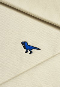 Tessuto beige con una trama testurizzata, caratterizzato da un design di un dinosauro ricamato in blu. I punti di cucitura delineano il dinosauro in nero.