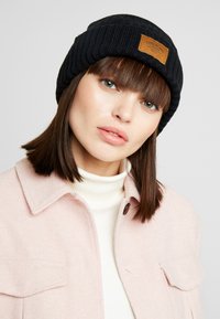 Mustahomea musta ribbed knit-pipo, jossa on vaaleanruskea nahkalogo, päällä henkilö, jolla on vaalea turtleneck-paita ja vaaleanpunainen takki.