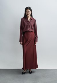 Robe en satin bordeaux avec manches longues, devant boutonné, et silhouette fluide ; associée à des chaussures plates noires. Fond uni.