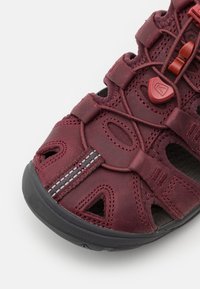 Maronen Sandale mit offenem Design, strukturiertem Lederoberteil, schwarzer Gummisohle und elastischen Schnürsenkeln. Merkmale sind reflektierende Nähte und Belüftungsöffnungen.