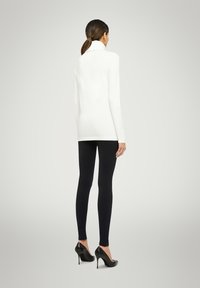 Weißes Langarmshirt kombiniert mit schwarzen Leggings und schwarzen High-Heels, von hinten vor einem hellgrauen Hintergrund gezeigt.