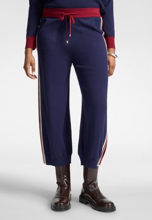Personne portant un pantalon bleu marine avec des bandes latérales rouges et blanches, une ceinture rouge, associé à des bottes en cuir marron foncé sur un fond uni.