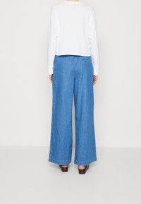 Weite blaue Jeanshosen mit hoher Taille, weicher Textur und subtilem Steppdetail. Kombiniert mit einem verkürzten weißen Langarmoberteil.