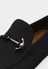 Mocassin en cuir noir avec des coutures détaillées et un ornement en métal argenté en forme d'ancre sur la partie supérieure.