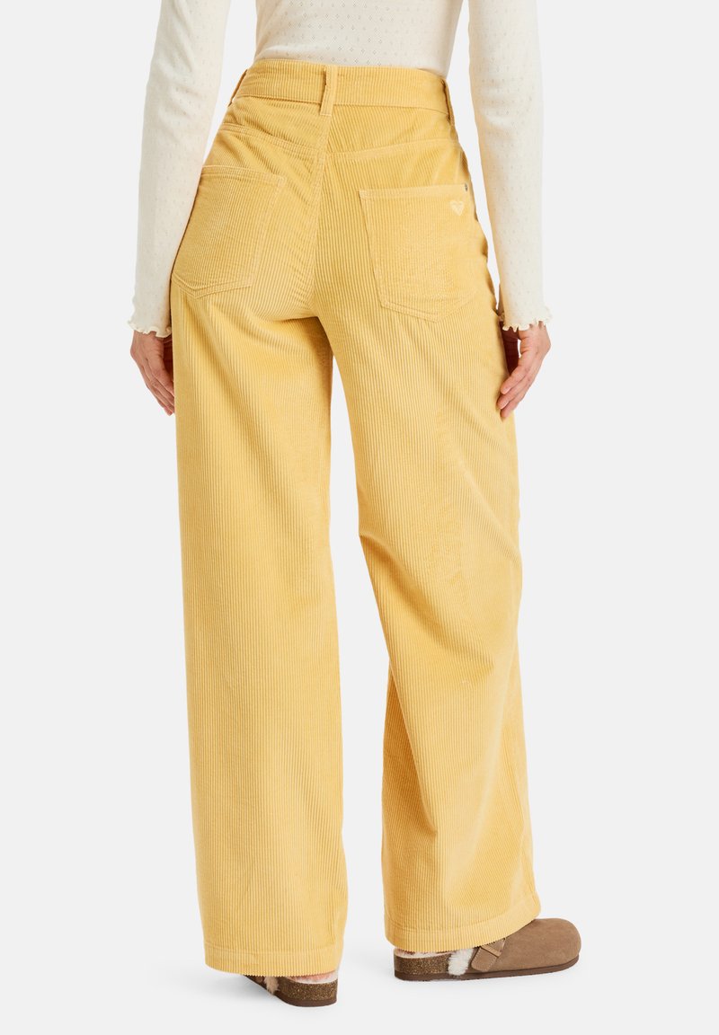 Pantalons larges en velours côtelé jaune avec poches arrière, présentant une texture côtelée verticale et une taille haute, complétés par des chaussures à enfiler marron.