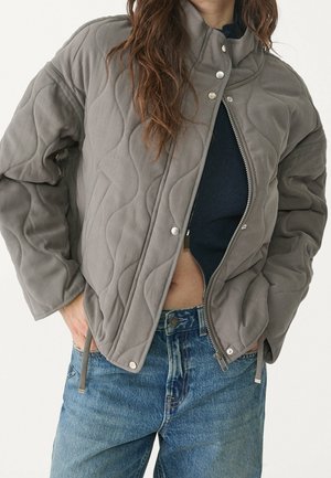 Frau trägt eine graue gesteppte Jacke über einem marineblauen Oberteil und blauen Jeans, die Hände in den Jackentaschen, die Jacke teilweise geöffnet.