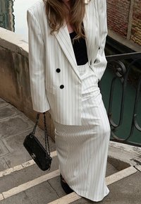 Blazer blanc à rayures et jupe assortie en tissu fluide. Comprend des boutons noirs, porté avec un haut noir et un sac à main noir.