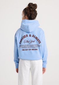 Donna con capelli ricci raccolti a chignon, che indossa un hoodie azzurro chiaro con la scritta "Forever & Always New York" e pantaloni bianchi, di spalle rispetto alla fotocamera.