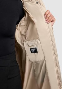 Beige gewatteerde jas met een gladde textuur en drukknoopsluiting. Het interieur bevat een patchzak en een merklabel.
