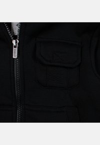 Veste en tissu noir avec une fermeture éclair argentée, présentant une petite poche à rabat avec des détails cousus sur la partie gauche de la poitrine.