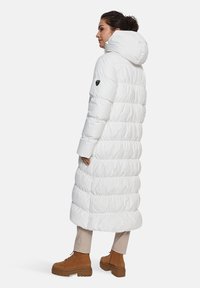 Witte lange pufferjas met een capuchon, met een gequilt ontwerp, een gladde textuur en een zwart logo op de mouw, gecombineerd met tan laarzen.
