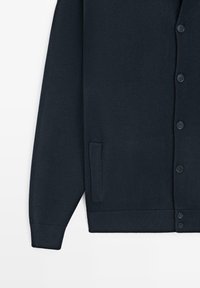 Marineblauer Strickcardigan mit geripptem Kragen, Bündchen und Saum. Er verfügt über sechs passende Knöpfe und eine seitliche Tasche auf der linken Seite.