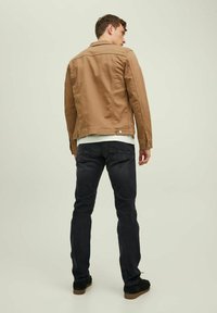 Chaqueta de algodón marrón con cuello y cierre de botones, combinada con jeans oscuros y zapatos negros. Presenta un corte ajustado y líneas limpias.