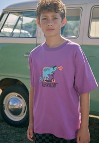 Paarse katoenen t-shirt met een chameleonafbeelding die op een scooter rijdt, vergezeld van een speelse draai en de tekst "HOU DOOR" eronder.