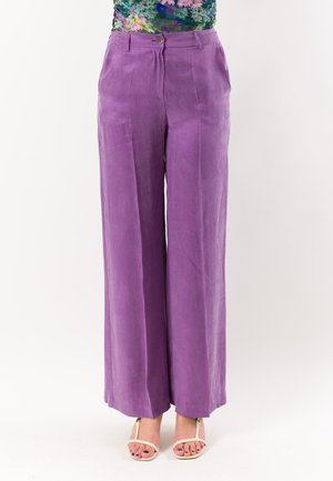 Donna che indossa pantaloni viola a vita alta e gamba larga e décolleté beige con tacco aperto su sfondo bianco semplice.