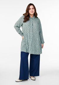 Long-sleeve, button-up shirt in soft green with a floral pattern, paired with wide-leg dark denim pants and light-colored shoes.
