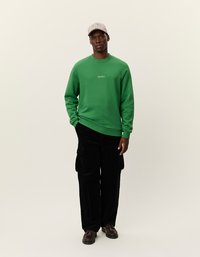 Žalios spalvos megztinis su užrašu "LES DEUX", derinamas su juodomis kargo kelnėmis ir rudomis bateliais. Modelis vilki dryžuotą kepurę. Lygus fonas.