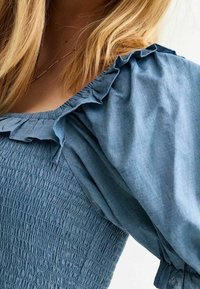 Světle modrá blůzka s volánkovými rukávy a nabíraným živůtkem. Vyznačuje se měkkou strukturou látky a volným střihem, který zdůrazňuje pohodlí a styl.