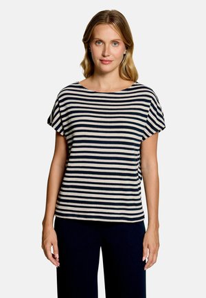 Femme aux cheveux blonds portant un haut à manches courtes rayé horizontalement noir et blanc et un pantalon noir, debout devant un fond clair uni.