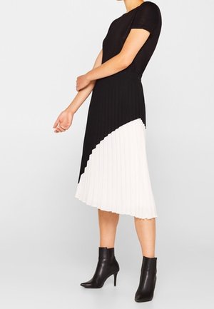 Vrouw die een zwart-witte geplooide midi-jurk met korte mouwen en zwarte enkellaarsjes met hakken draagt, staand tegen een effen achtergrond.