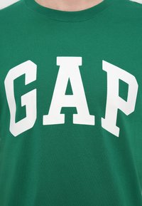 Πράσινο κοντομάνικο βαμβακερό μπλουζάκι με μεγάλα λευκά γράμματα "GAP". Ομαλή υφή με στρογγυλή λαιμόκοψη.