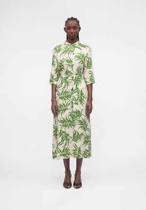 Femme debout portant une longue robe crème à motifs de feuilles vertes, manches 3/4, taille ceinturée et talons ouverts marron, sur fond blanc uni.