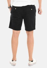 Schwarze Baumwollshorts mit geradem Schnitt, ausgestattet mit zwei Knöpfen an den hinteren Taschen. Getragen mit weißen Turnschuhen und einem einfachen weißen T-Shirt.