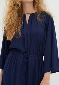 Žena s dlhými kučeravými blond vlasmi, oblečená v voľných tmavomodrých šatách so šnúrkou v páse a výstrihom v tvare kľúčovej dierky.