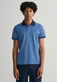 Modrá polo tričko se strukturovanou tkaninou, tmavým límečkem a pruhovanými akcenty na rukávech. Obsahuje malé logo na levé hrudi.