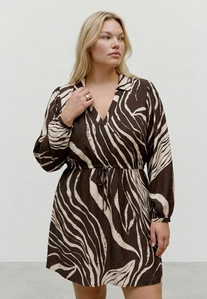 Femme aux cheveux blonds portant une robe zébrée marron et beige à manches longues avec une ceinture à nouer, debout devant un fond clair uni.