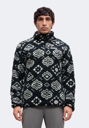Ung mand iført en sort og hvid mønstret Columbia fleece-trøje med kvart lynlås og grå bukser, stående mod en ensfarvet baggrund.
