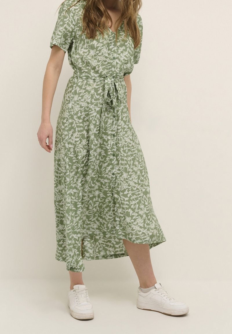 Femme portant une robe midi verte à fleurs avec des boutons et une ceinture nouée à la taille, associée à des baskets blanches, debout sur un fond clair.