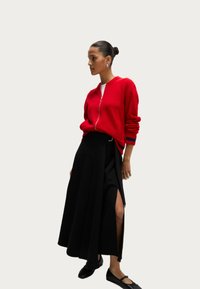 Cardigan rosso con zip e dettagli a maglia, abbinato a una gonna nera a vita alta con spacco laterale e scarpe nere flat.
