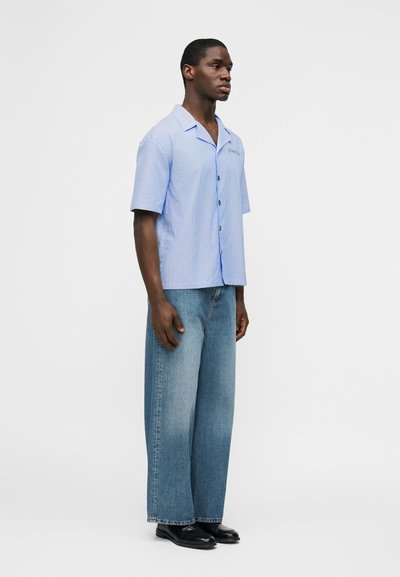 Νεαρός άνδρας που στέκεται πλάγια φοράει ανοιχτό μπλε πουκάμισο με κοντά μανίκια και κουμπιά, φαρδιά ξεθωριασμένα μπλε τζιν και μαύρα παπούτσια.