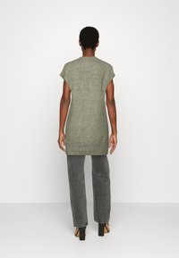 Tunic verde a maglia con maniche corte, finitura testurizzata, scollatura alta e spacchi laterali, abbinata a pantaloni grigi larghi e sandali con tacco a blocco.
