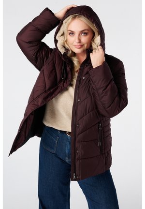 Jeune femme portant une veste matelassée à capuche marron foncé sur un pull beige et un jean bleu, ajustant la capuche de la veste d'une main.