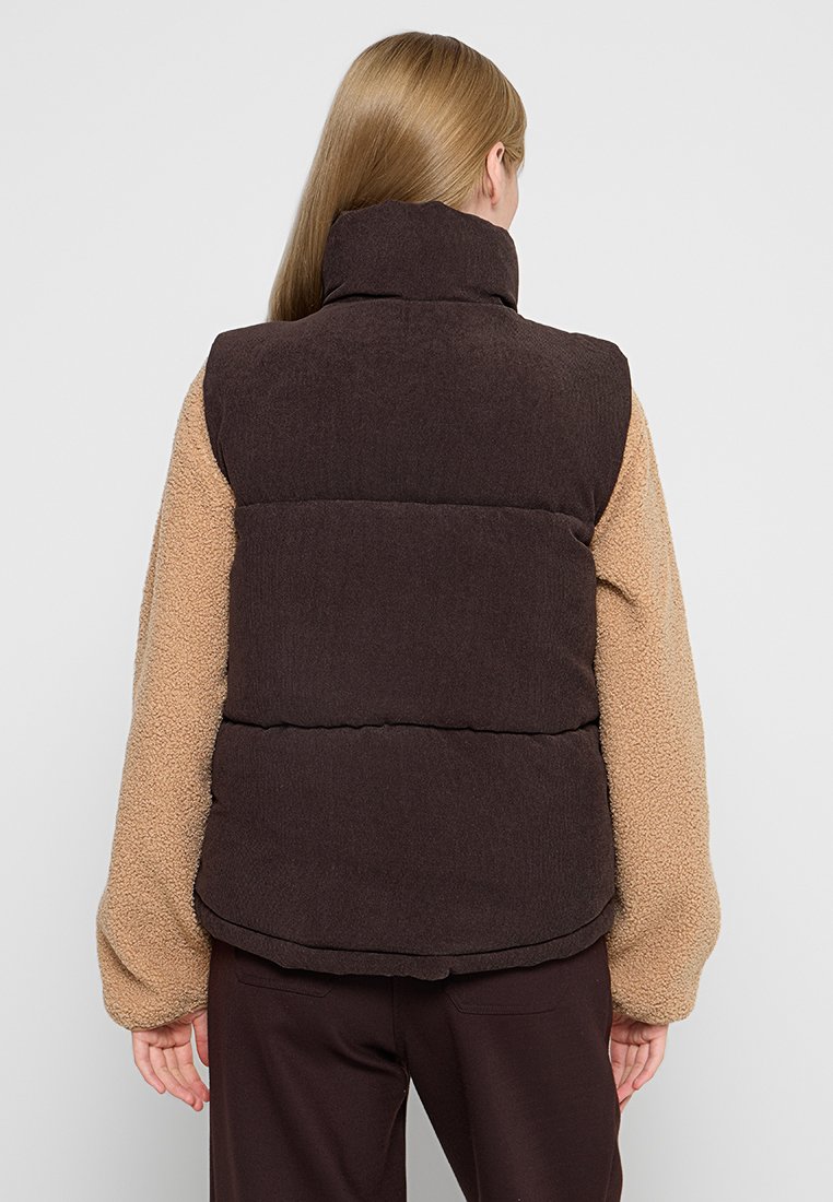 Braune Steppweste mit hohem Kragen und plüschigen, beigen Ärmeln. Sie verfügt über einen strukturierten Stoff und eine taillierte Taille für eine figurbetonte Passform.