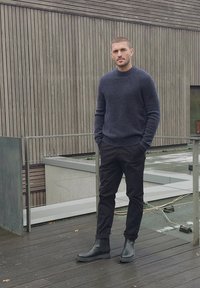 Homme portant un pull foncé, un pantalon noir et des bottes noires, debout sur une terrasse en bois près d'un bâtiment moderne avec un bardage en bois vertical.